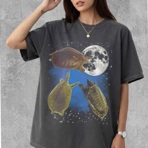 Może przedstawiać: Ciemnoszary t-shirt z krótkim rękawem z nadrukiem graficznym przedstawiającym trzy żółwie pływające na tle ciemnoniebieskiego rozgwieżdżonego nieba, z dużym, szczegółowym białym księżycem w prawym górnym rogu wzoru.