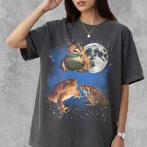 May include: Dark gray t-shirt featuring a whimsical design of three toads against a blue and starry night sky, with a full moon in the background. The top toad has its tongue out. A unique and fun graphic tee.
