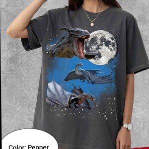 May include: Dark gray t-shirt featuring a fantasy design with three dragons, a full moon, and a starry sky. The dragons are in shades of gray and blue, with one dragon's mouth open. The shirt has a crew neck and short sleeves.