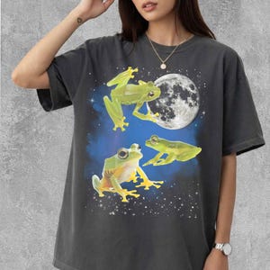 Può includere: T-shirt grigio scuro con un design stravagante di tre rane verdi su uno sfondo di luna piena e cielo stellato. Le rane sono raffigurate in varie pose, aggiungendo un tocco giocoso al design.