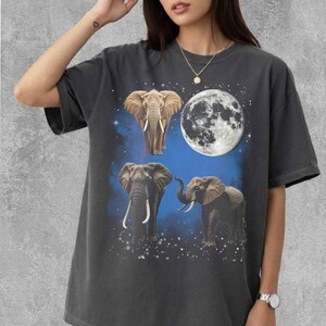 May include: Dark grey t-shirt with a graphic of three elephants, a full moon, and a blue starry background. The elephants are brown and grey, with long white tusks. The moon is white with detailed craters, creating a celestial theme.