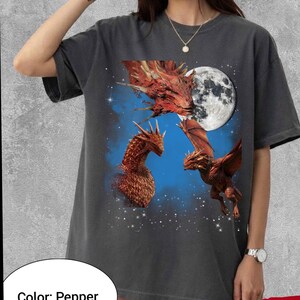 May include: Pepper-colored t-shirt featuring a fantasy design. The graphic depicts three red dragons against a blue and starry background, with a full moon. The dragons have detailed scales and wings.