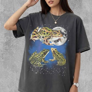Può includere: T-shirt grigio scuro con un design a rana stravagante. La grafica include tre rane, una luna piena e un cielo stellato. Le rane sono verdi, gialle e marroni con un motivo maculato. La maglietta è uno stile casual e unisex.