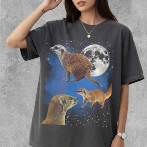 May include: Dark gray t-shirt featuring a graphic of three meerkats against a backdrop of a full moon and a starry night sky. The meerkats are depicted in shades of brown and tan.