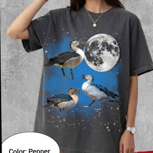 May include: Pepper-colored t-shirt featuring a graphic design of three ducks, a full moon, and stars. The ducks have black, white, and brown feathers. The background is blue with white stars. The shirt has short sleeves.
