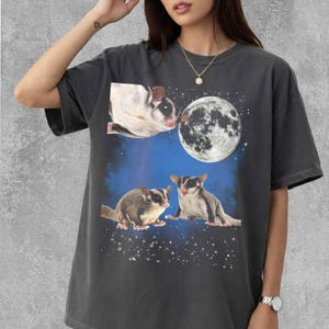 Puede incluir: Camiseta gris oscuro con un gráfico de tres petauros del azúcar, una luna llena y un cielo estrellado. El diseño incluye un petauro del azúcar cerca de la luna y dos petauros debajo. La camiseta es de estilo casual y unisex.