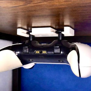 Könnte beinhalten: Ein schwarz-weißer Gamecontroller, der von einer weißen Halterung gehalten wird, die an einer Holzoberfläche befestigt ist. Der Controller ist für Spiele konzipiert und hat ein schlankes, ergonomisches Design.