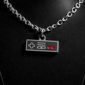 May include: A silver-toned metal chain necklace featuring a rectangular pendant shaped like a retro video game controller. The pendant is grey with a black center, a white directional cross, white horizontal lines, and two circular red buttons.