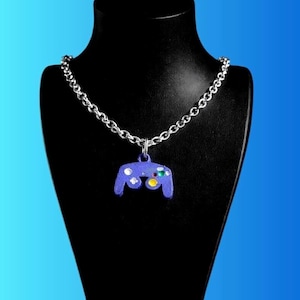 Puede incluir: Collar de cadena plateada con un colgante con forma de mando de videojuego morado. El mando tiene botones de colores y se muestra sobre un maniquí negro sobre un fondo degradado azul.