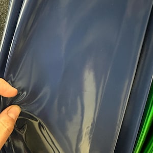 May include: A close-up of a roll of navy blue vinyl material. The material has a glossy finish and is slightly creased. The vinyl is being held by a hand.