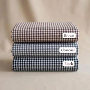 May include: Three folded stacks of fabric featuring a small gingham check pattern. The top stack is brown and white, the middle stack is charcoal and white, and the bottom stack is black and white. Each stack has a white label with the color name.
