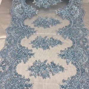 May include: A light blue lace fabric featuring intricate floral embroidery and scalloped edges. The design is embellished with small, shimmering beads and sequins arranged in repeating patterns across the sheer, lightweight mesh material.