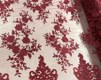 May include: Burgundy lace fabric with floral and ornamental patterns. The fabric features detailed embroidery of roses and leaves, creating a textured design. The lace is semi-sheer, with a rolled edge visible on the right side. Ideal for dresses, veils, or decorative projects.