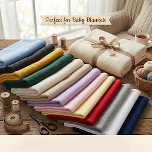 Pode incluir: Uma coleção de mantas macias para bebês em várias cores, incluindo azul, cinza, amarelo, verde, bege e vermelho, dispostas em uma superfície de madeira. Uma manta creme é amarrada com uma fita. Suprimentos de costura estão presentes.