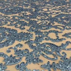 Puede incluir: Primer plano de una tela de encaje azul marino con lentejuelas. La tela presenta un intrincado diseño floral y de volutas, con lentejuelas que añaden un efecto brillante. El encaje se coloca sobre un fondo neutro, resaltando el patrón detallado.