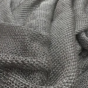 Könnte beinhalten: Nahaufnahme eines drapierten, metallisch silbernen Kettenstoffes. Das Material ist dicht gewebt und erzeugt eine strukturierte Oberfläche mit dezentem Glanz. Der Stoff ist gefaltet und zeigt Tiefe und Dimension.