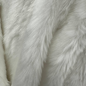 May include: Close-up of a white faux fur fabric. The fabric has a soft, fluffy texture with long, flowing fibers. The fibers are a bright white color, creating a luxurious appearance. The fabric is likely used for clothing or home decor.