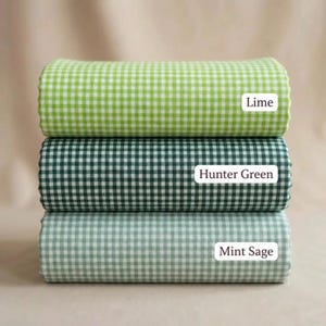 Puede incluir: Tres rollos de tela apilados con un patrón de cuadros vichy. El rollo superior está etiquetado como Lime, el central como Hunter Green y el inferior como Mint Sage. Cada rollo muestra una cuadrícula consistente de color blanco y verde.