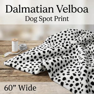 May include: Dalmatian Velboa fabric with a dog spot print, 60 inches wide. The white fabric is covered in black spots. A spool of white thread, a measuring tape, and a small measuring tape are on the wooden surface.