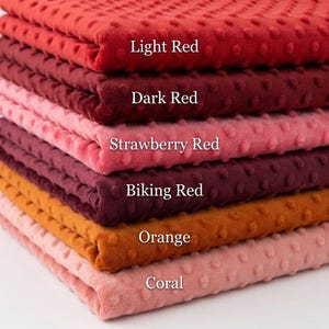 May include: A stack of textured fabric samples in shades of red, orange, and coral. The colors are labeled "Light Red", "Dark Red", "Strawberry Red", "Biking Red", "Orange", and "Coral". The fabric has a dotted pattern.