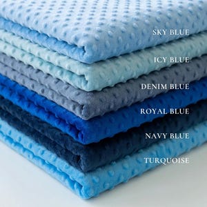 Op de afbeelding: Een stapel zachte, getextureerde dekens in verschillende blauwtinten, waaronder hemelsblauw, ijsblauw, spijkerbroekblauw, koningsblauw, marineblauw en turkoois. Elke deken heeft een gestippeld patroon en is voorzien van de kleurnaam.