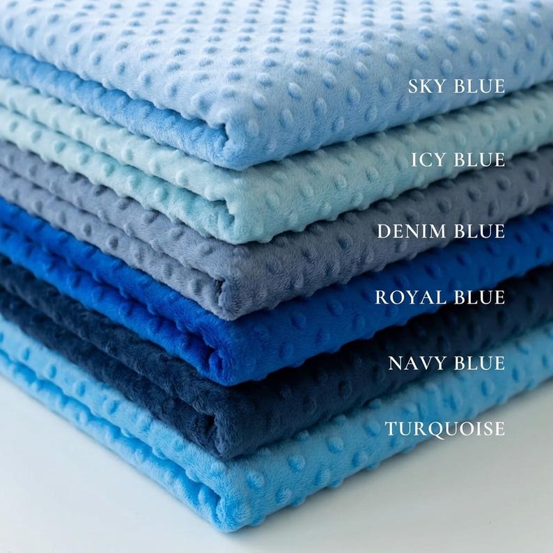 May include: A stack of soft, textured blankets in various shades of blue, including sky blue, icy blue, denim blue, royal blue, navy blue, and turquoise. Each blanket has a dotted pattern and is labeled with its color name.