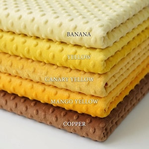 Könnte beinhalten: Ein Stapel Plüschstoff in Gelb- und Brauntönen. Die Farben umfassen Banane, Gelb, Kanariengelb, Mangogelb und Kupfer. Jeder Stoff hat eine strukturierte, gepunktete Oberfläche. Die Farbbezeichnungen sind auf dem Stoff aufgedruckt.