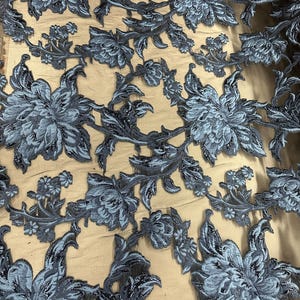 Könnte beinhalten: Nahaufnahme eines Stoffes mit floralem Muster. Das Design zeigt große, detaillierte Blumen und Blätter in Blau- und Grautönen, wodurch ein strukturiertes, dreidimensionales Aussehen entsteht. Der Stoff scheint Spitze oder ein besticktes Material zu sein, geeignet für Kleidung oder Dekorationsprojekte.