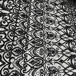 May include: Close-up of black embroidered lace fabric with a detailed floral and scrollwork pattern. The lace is set against a white background, creating a high-contrast, elegant design. The fabric is likely used for apparel or home decor.