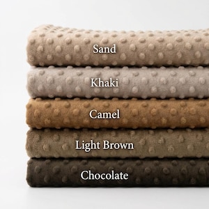 Könnte beinhalten: Ein Stapel von fünf Plüschdecken in verschiedenen Brauntönen. Die Farben sind mit "Sand", "Khaki", "Camel", "Light Brown" und "Chocolate" beschriftet. Jede Decke hat eine strukturierte, gepunktete Oberfläche.