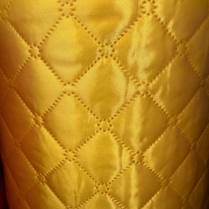 May include: Close-up of a quilted fabric in a vibrant golden yellow color. The fabric features a diamond pattern created by stitched lines. The texture appears smooth and slightly shiny, suggesting a satin or silk material. The quilting adds depth and visual interest.
