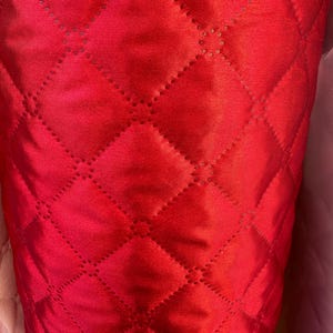 May include: Close-up of a bright red quilted fabric. The fabric features a diamond pattern formed by stitched lines. The material has a subtle sheen, possibly satin or silk. The stitching is visible, adding detail to the design.