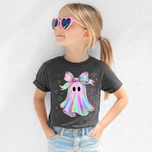 Puede incluir: Una camiseta gris oscuro de manga corta con un gr&aacute;fico central de un fantasma con un gran lazo iridiscente. El dise&ntilde;o del fantasma muestra una paleta de colores arco&iacute;ris met&aacute;licos, acentuada por peque&ntilde;as estrellas y corazones de colores.