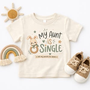 Puede incluir: Una camiseta de manga corta color crema con el gráfico de un conejo de dibujos animados sosteniendo un anillo de madera. El texto "My Aunt is SINGLE" y "ask my parents for details" aparece en letras verde oscuro y marrón, con estrellas y corazones.