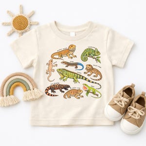 Peut inclure: Un t-shirt à manches courtes de couleur crème avec une impression graphique colorée représentant divers reptiles, dont un dragon barbu, un caméléon, un gecko, un scinque, un iguane, un lézard à collerette, un monstre de Gila, un lézard cornu et un anolis, chacun étiqueté avec son nom.