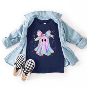 Puede incluir: Una camiseta azul marino oscuro con un gr&aacute;fico central de un fantasma hologr&aacute;fico que lleva un gran lazo iridiscente, rodeado de peque&ntilde;as estrellas y corazones de colores, junto a una camisa vaquera azul claro y zapatillas de cuadros.