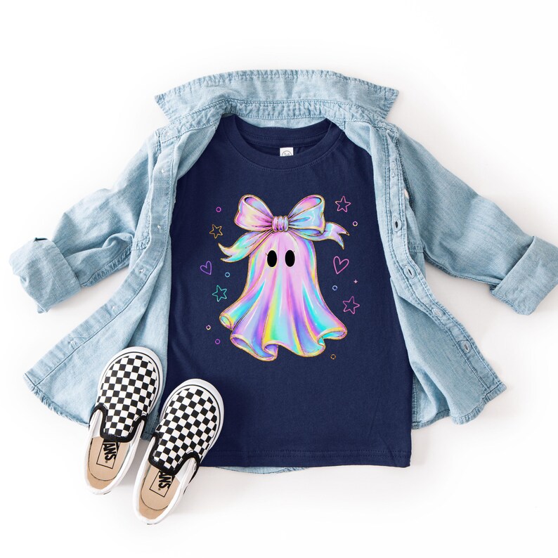 Puede incluir: Una camiseta azul marino oscuro con un gr&aacute;fico central de un fantasma hologr&aacute;fico que lleva un gran lazo iridiscente, rodeado de peque&ntilde;as estrellas y corazones de colores, junto a una camisa vaquera azul claro y zapatillas de cuadros.