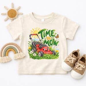 Pode incluir: Uma t-shirt de manga curta cor de creme com um gráfico de um corta-relvas vermelho sorridente em relva verde com dentes-de-leão amarelos e um sol. O texto "TIME TO MOW" está escrito em letras verdes. Inclui uma pequena borboleta laranja.