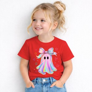 Puede incluir: Una camiseta roja de manga corta con un gr&aacute;fico central de un fantasma que lleva un lazo grande, iridiscente y multicolor. El dise&ntilde;o del fantasma incluye tonos pastel suaves de rosa, azul y amarillo, rodeado de peque&ntilde;as estrellas y corazones.