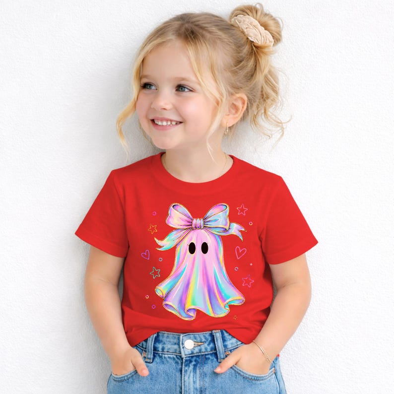 Puede incluir: Una camiseta roja de manga corta con un gr&aacute;fico central de un fantasma que lleva un lazo grande, iridiscente y multicolor. El dise&ntilde;o del fantasma incluye tonos pastel suaves de rosa, azul y amarillo, rodeado de peque&ntilde;as estrellas y corazones.