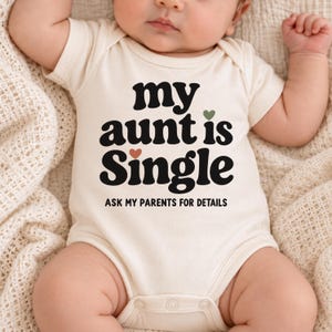 Puede incluir: Un body para bebé de color crema con el texto negro "my aunt is Single", un pequeño corazón naranja y un pequeño corazón verde, seguido del texto "ASK MY PARENTS FOR DETAILS" en una fuente negra más pequeña en el área del pecho.
