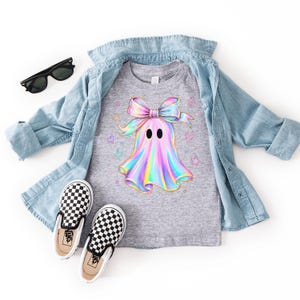 Puede incluir: Una camiseta gris jaspeado con un gr&aacute;fico de fantasma hologr&aacute;fico con un lazo grande, rodeado de peque&ntilde;as estrellas y corazones de colores. La camiseta se lleva bajo una camisa vaquera azul claro, junto con gafas de sol negras y zapatillas a cuadros.