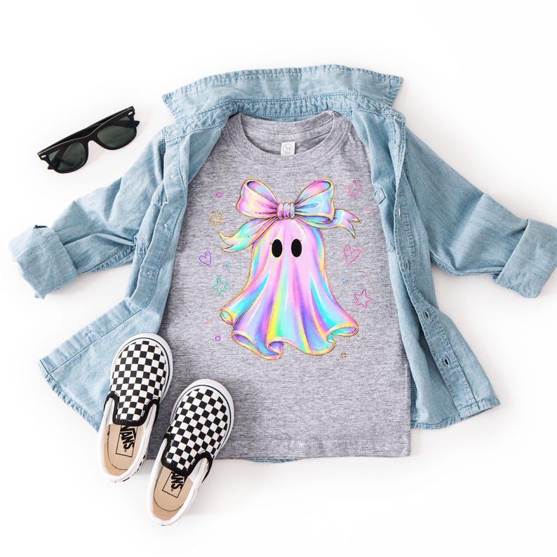 Puede incluir: Una camiseta gris jaspeado con un gr&aacute;fico de fantasma hologr&aacute;fico con un lazo grande, rodeado de peque&ntilde;as estrellas y corazones de colores. La camiseta se lleva bajo una camisa vaquera azul claro, junto con gafas de sol negras y zapatillas a cuadros.