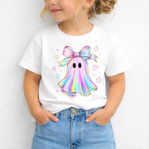 Puede incluir: Una camiseta blanca de manga corta con un gr&aacute;fico central de un fantasma con un gran lazo iridiscente en la cabeza. El fantasma tiene un acabado met&aacute;lico de colores arco&iacute;ris con dos ojos ovalados negros, rodeado de peque&ntilde;as estrellas y corazones.