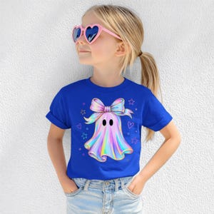 Puede incluir: Una camiseta de manga corta azul real con un gr&aacute;fico central de un fantasma de estilo hologr&aacute;fico que lleva un lazo grande, rodeado de peque&ntilde;as estrellas y corazones delineados. El fantasma tiene dos ojos ovalados negros y un acabado iridiscente.