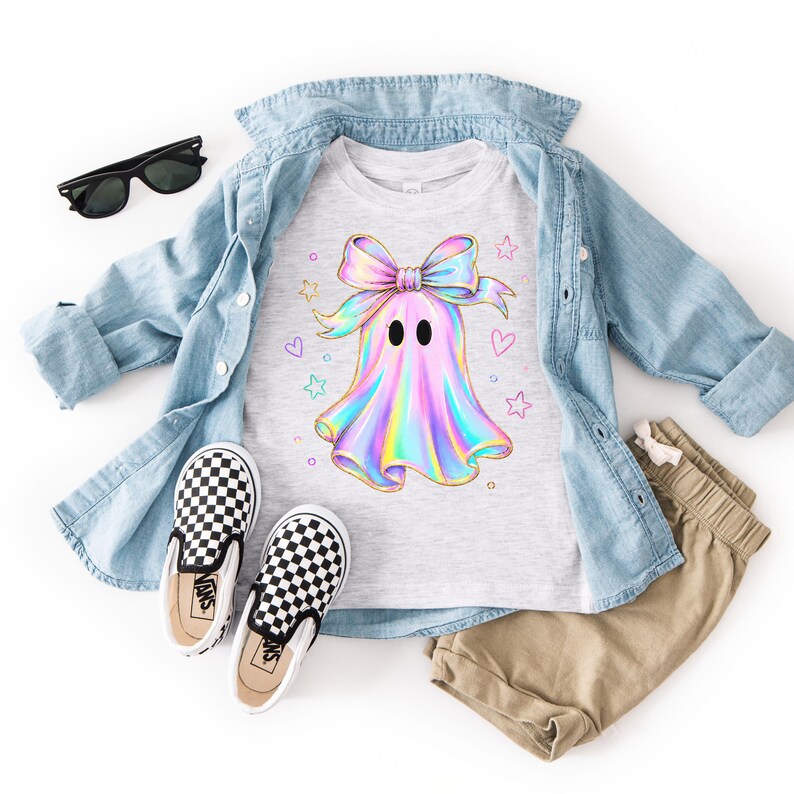 Puede incluir: Una camiseta gris claro con el gr&aacute;fico de un fantasma de estilo hologr&aacute;fico con un lazo grande, rodeado de estrellas y corazones peque&ntilde;os. La prenda se muestra con una camisa azul claro, pantalones cortos beige, gafas de sol negras y zapatos a cuadros.