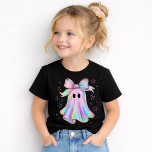 Puede incluir: Una camiseta negra de manga corta con un gr&aacute;fico central de un fantasma con un gran lazo iridiscente en la cabeza. El fantasma tiene un acabado met&aacute;lico de colores arco&iacute;ris y est&aacute; rodeado de peque&ntilde;os contornos de estrellas y corazones.