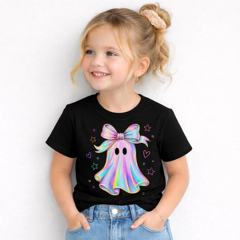 Puede incluir: Una camiseta negra de manga corta con un gr&aacute;fico central de un fantasma con un gran lazo iridiscente en la cabeza. El fantasma tiene un acabado met&aacute;lico de colores arco&iacute;ris y est&aacute; rodeado de peque&ntilde;os contornos de estrellas y corazones.