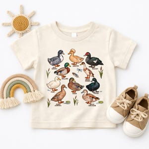 Peut inclure: Un t-shirt à manches courtes de couleur crème avec une impression graphique colorée représentant diverses races de canards, notamment Blue Swedish, Crested, Muscovy, Mallard, Indian Runner, Pekin, Rouen, Cayuga, Khaki Campbell et Welsh Harlequin.