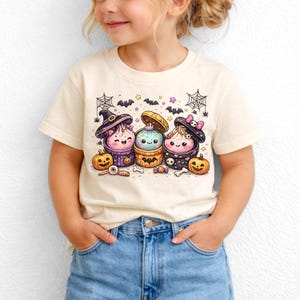 Peut inclure: Un t-shirt couleur crème avec une impression graphique représentant trois personnages ronds et mignons dans des récipients en forme de citrouille. Le motif inclut des petites chauves-souris, des toiles d'araignée, des étoiles, des citrouilles orange, un crâne et des bonbons, le tout dans un style cartoon coloré.